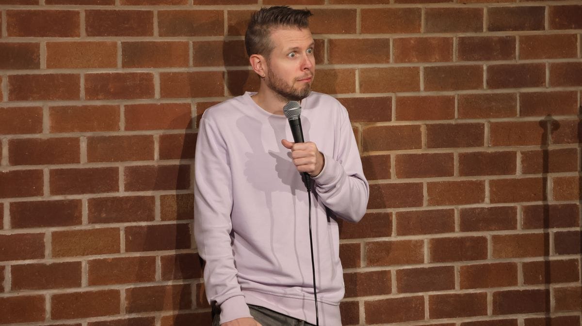 Phil Chapman on stage against a brick wall, eyebrows raised and lips pursed, microphone at his chest