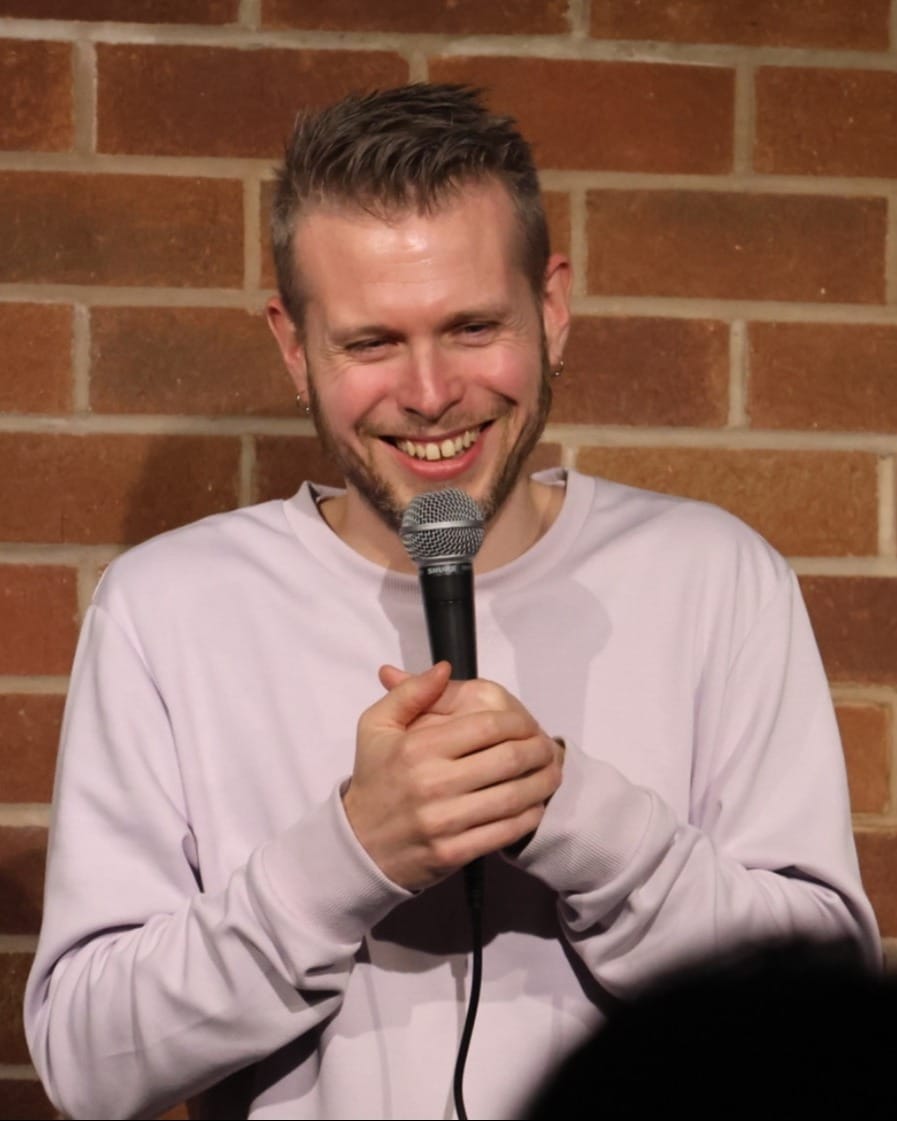 Phil Chapman laughing on stage, microphone in both hands, in front of a brick wall