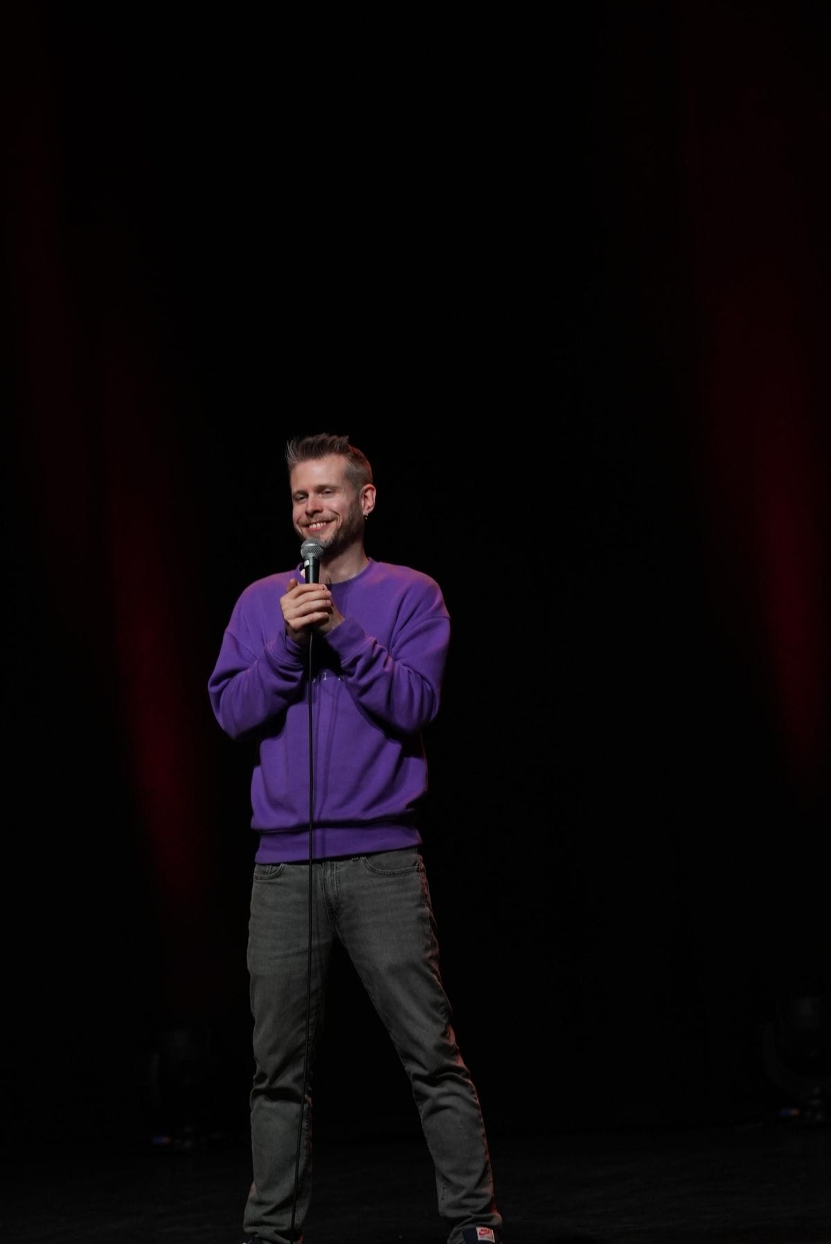 Phil Chapman alone on a dark stage in a purple jumper, smiling into the microphone, a red curtain behind him