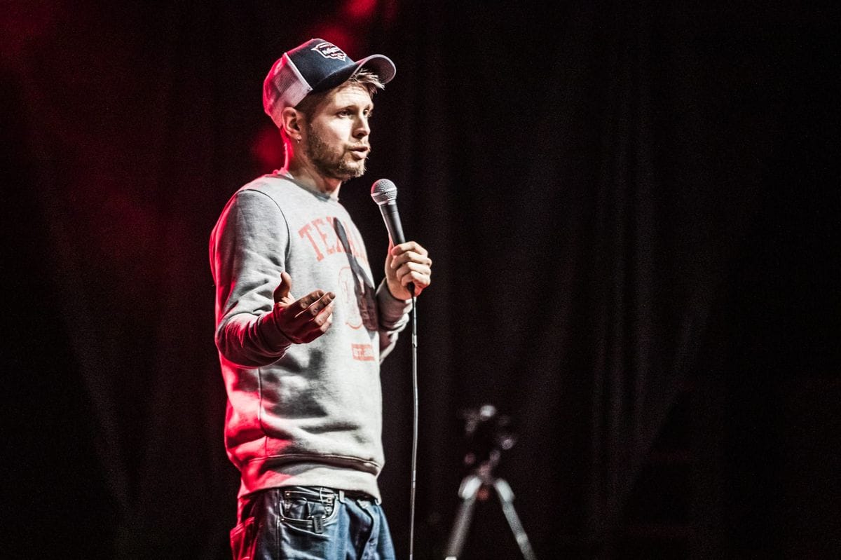 Phil Chapman performing under red stage light in a cap and Texans sweatshirt, mid-gesture with the microphone